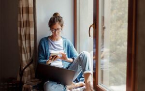 Woman sitting near window with smartphone and laptop
