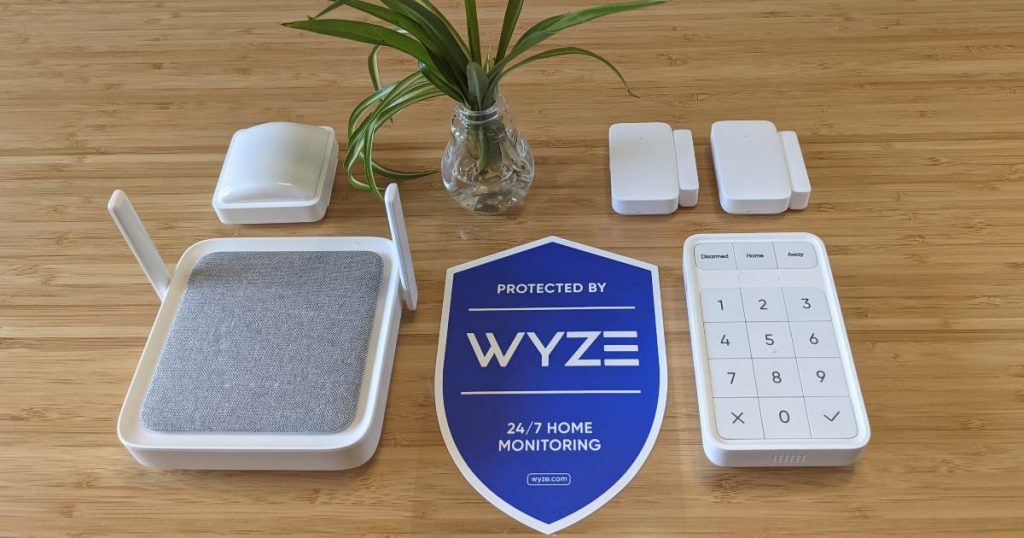 Shows a Wyze home security system laying on a wooden table: A motion sensor, two contact sensors, the Wyze Sense Hub, a yard sign, and a key pad.