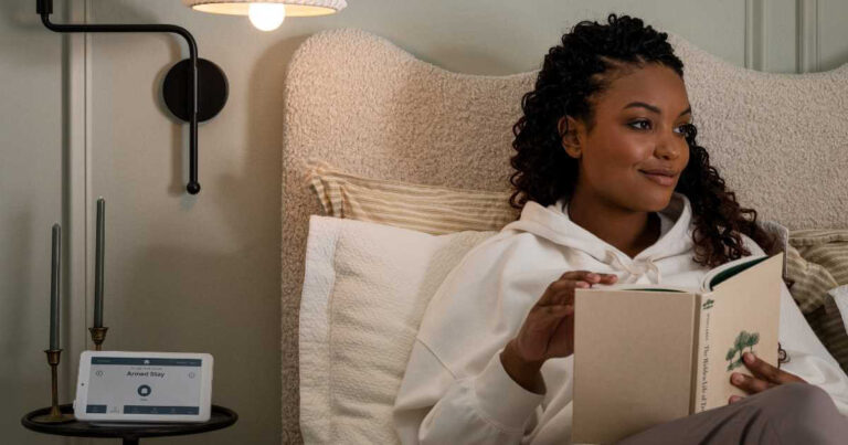 Woman reading in bed with Cove alarm panel on bedside table displaying "Armed Stay"