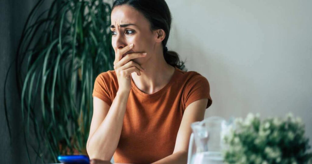 Sad, frustrated young brunette woman is crying with smartphone in hands while she sitting on the chair at apartment