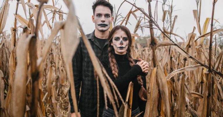 Spooky young couple in Halloween costumes in a cornfield standing one in front of the other and holding hands