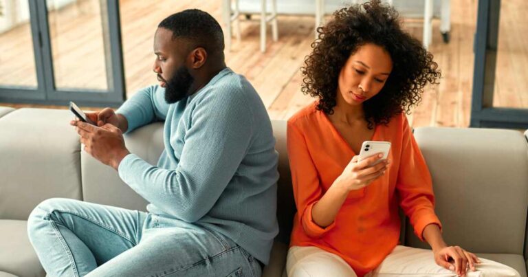 African American couple sitting on sofa with phones facing away from each other in living room at home.