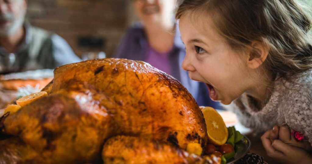 Cute little girl having fun while about to bite a stuffed turkey during Thanksgiving dinner in dining room.