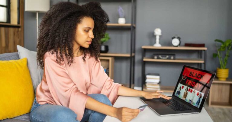 A multiracial woman is sitting on a couch, while typing on her laptop on the coffee table, holding a credit card in her hand as she completes her online purchase