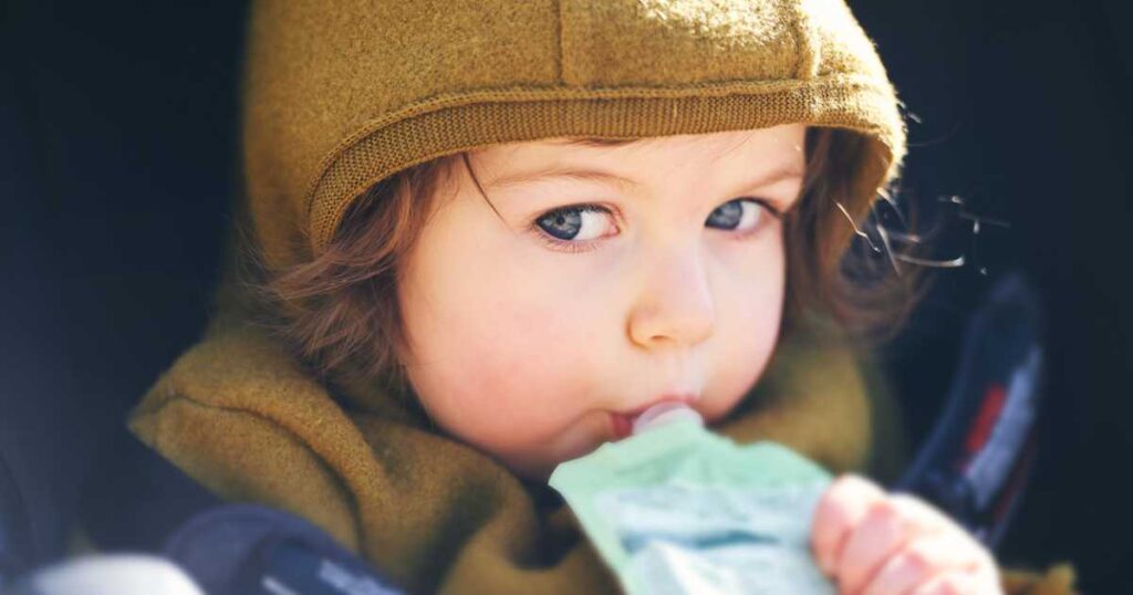 Close up portrait of sweet toddler kid eating fruit puree from plastic pouch, sitting in stroller, outdoor snack time.