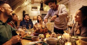 Multiracial group of friends gathering at dining table for Thanksgiving dinner. Focus is on man carving roast turkey.