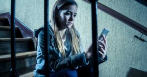Sad, depressed young teenage girl victim of cyberbullying by mobile smart phone sitting on stairs feeling lonely, unhappy, hopeless, and abused. Bullied by text message on social media app. Dark light