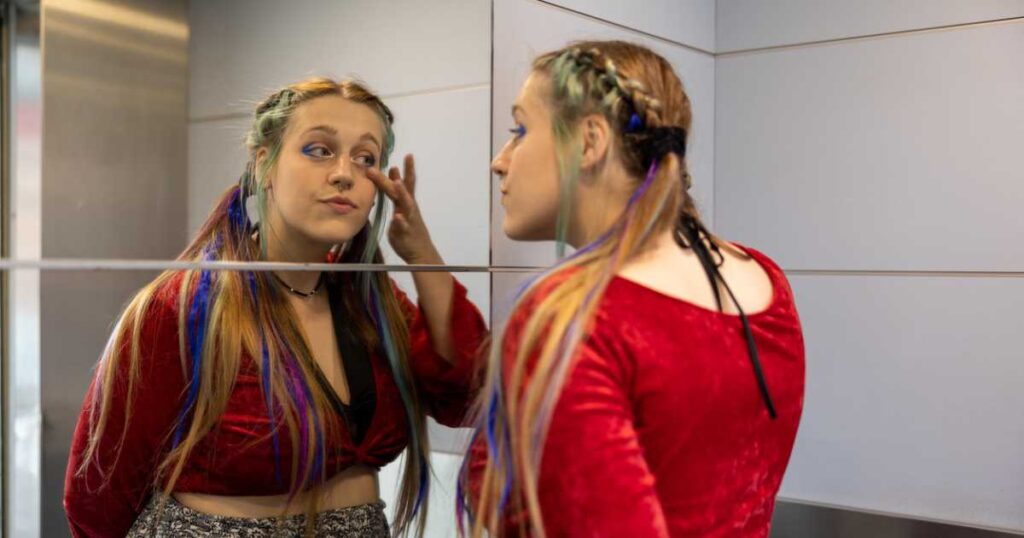 influencer girl checking herself in the mirror, wearing a red sweater with braided and dyed hair