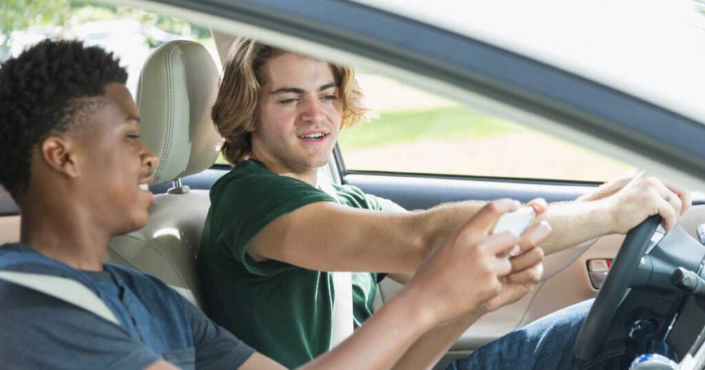 A caucasian teenage boy or young man sitting in the driver's seat of a car. His African American passenger is showing him something on his mobile phone.