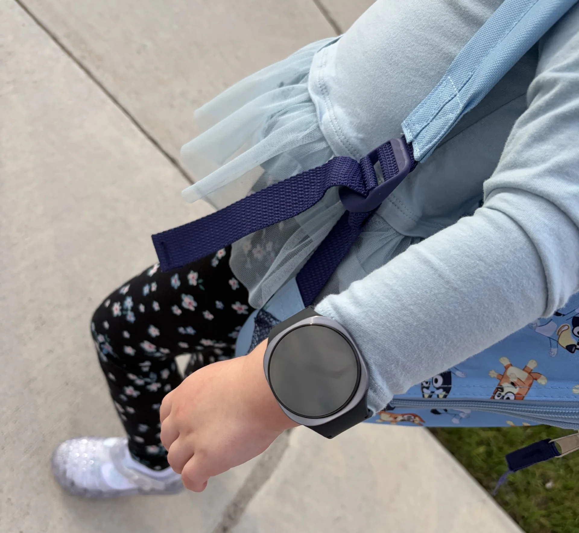 Child's wrist showing a Bark smartwatch with a dark round face and navy blue band, wearing light blue long sleeves and patterned socks while standing outdoors on pavement.