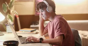 Side view portrait of teenage boy using laptop at night with wireless headphones in dimly lit room