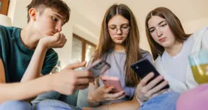 One teenage boy and two teenage girls looking at their mobile phones at home.