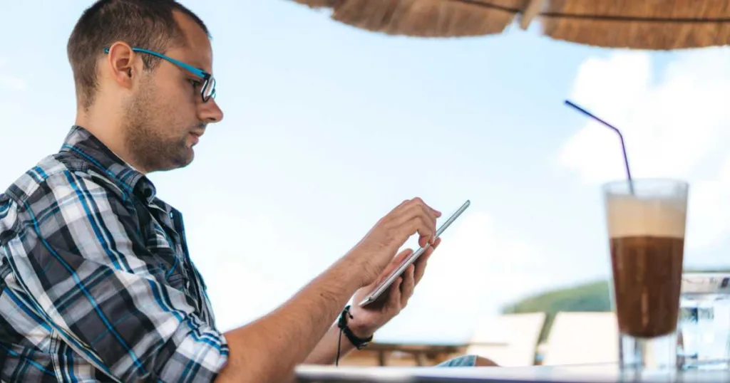 Young caucasian man checking in on his home on tablet and drinking ice coffee at seaside.