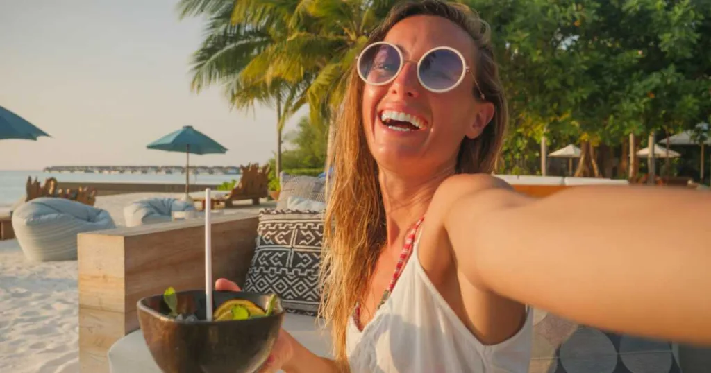 Woman having cocktail at sunset on tropical beach and taking selfie