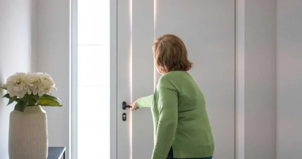 An older woman, wearing a green sweater and jeans, opens a bright door in a modern home interior with soft natural lighting and minimal decor.